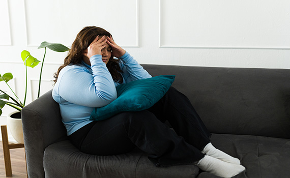Uma mulher triste e com excesso de peso, sentada num sofá, que sofre com os estigmas sociais associados ao peso. 