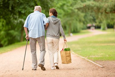 Um homem mais velho é ajudado a caminhar por uma mulher, que carrega as compras do idoso. 
