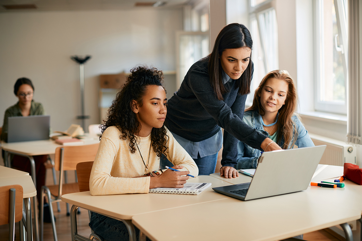 Em uma sala de aula, um professor de ensino médio auxilia dois alunos, dando-lhes instruções sobre o laptop. 