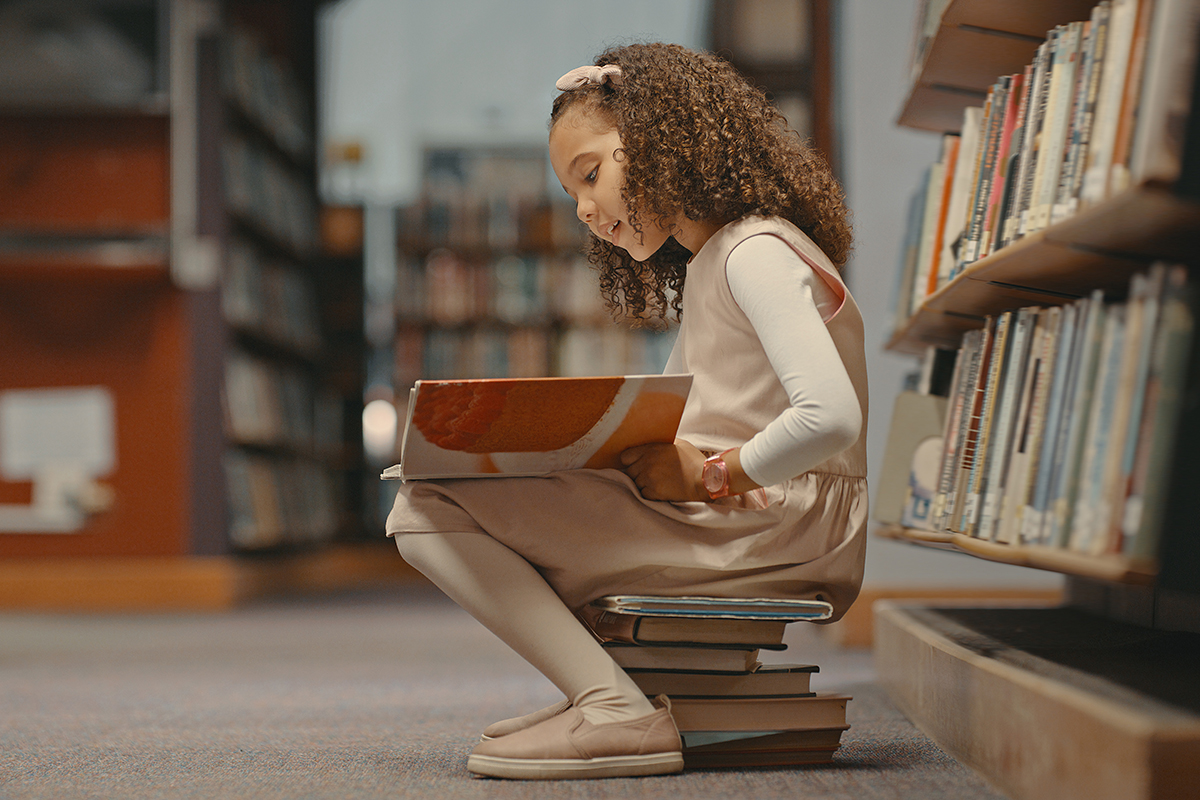 Menina sentada sobre livros na biblioteca e lendo um livro. 