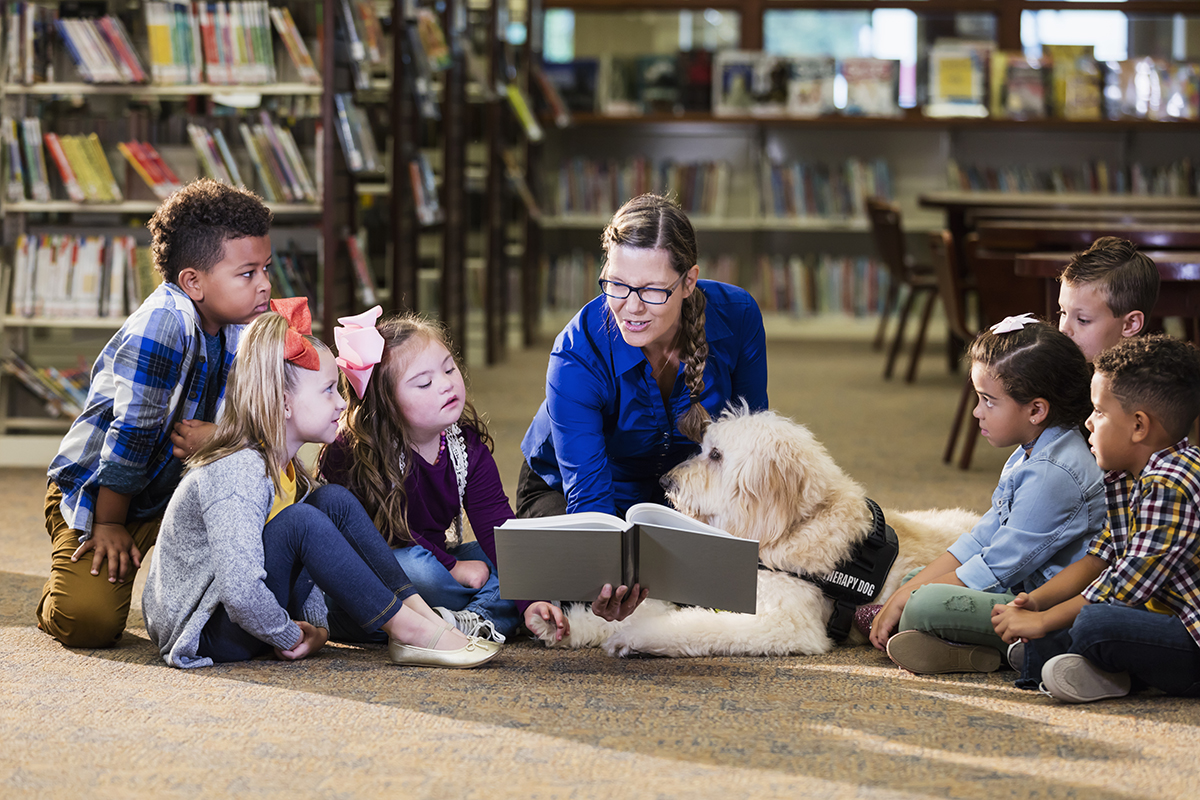 Sentados no chão da biblioteca, uma professora e seus alunos leem um livro, acompanhados por um cão de terapia. 