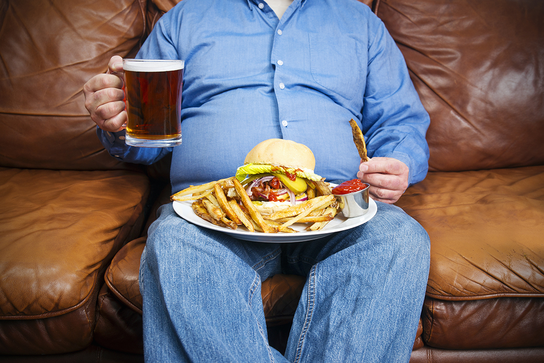 Pessoa obesa sentada em uma poltrona segurando uma cerveja e comendo um hambúrguer com batatas fritas. 