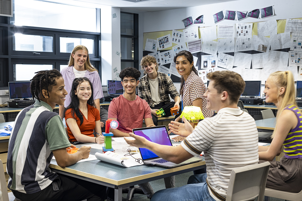 Foto grande de um grupo de alunos e seu professor reunidos em torno de uma mesa em uma sala de aula de tecnologia.
