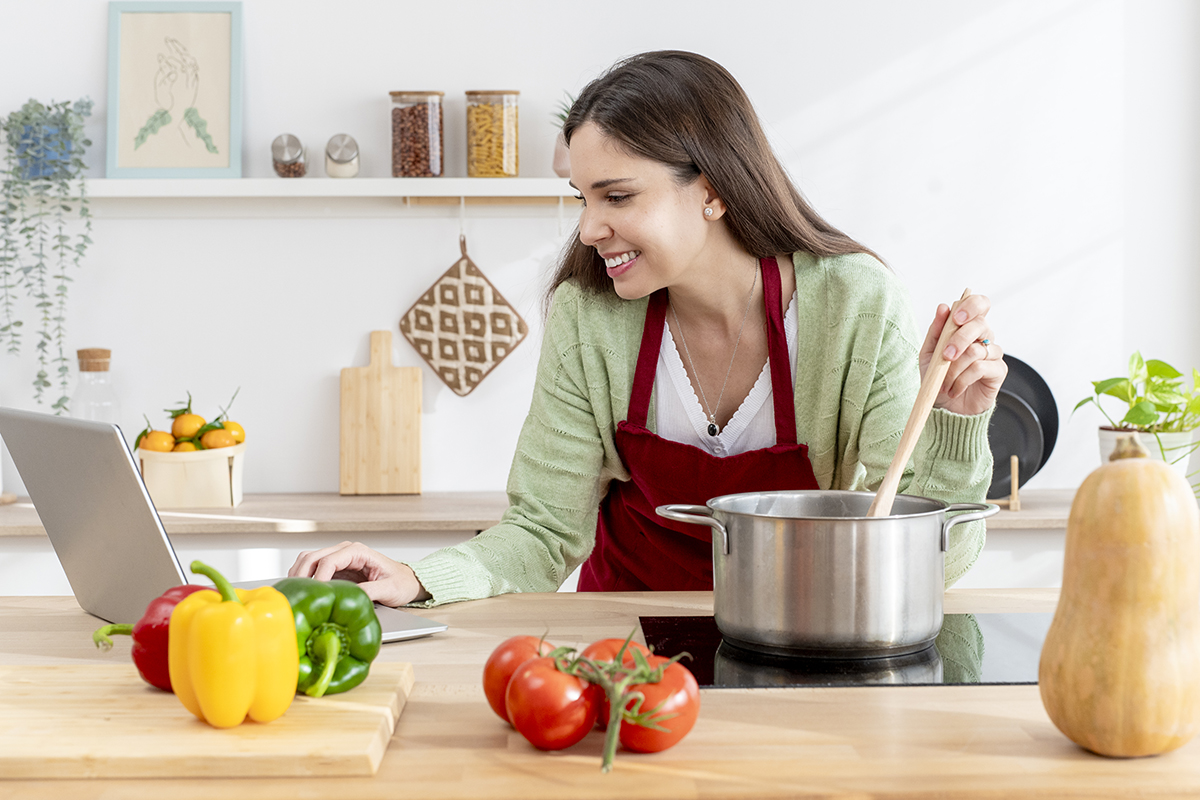 dona-de-casa-feliz-agitando-uma-panela-e-lendo-receita-em-um-laptop-cercado-de-vegetais-frescos-em-uma-cozinha-moderna.