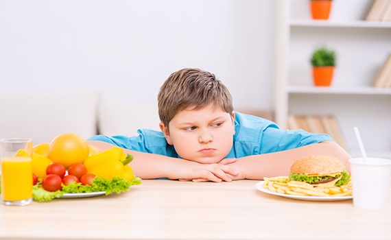 Un niño con sobrepeso viendo un plato de comida rápida, mientras ignora los alimentos sanos que tiene en frente. 
