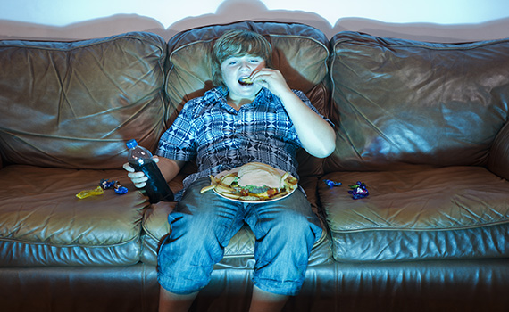 Un niño sentado en un sofá, viendo la televisión y comiendo alimentos ultraprocesados por la noche. 