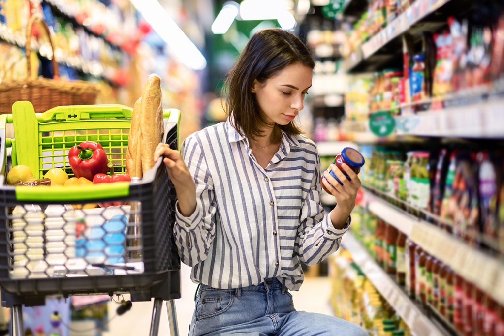 Mujer agachada en el pasillo del supermercado sostiene un bote mientras lee la etiqueta del producto. 