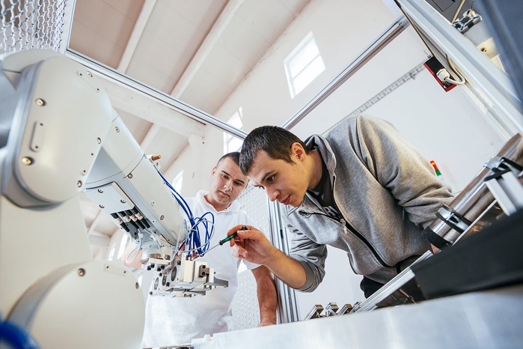 Dos personas realizan ajustes a una máquina que utiliza un brazo de robot. 