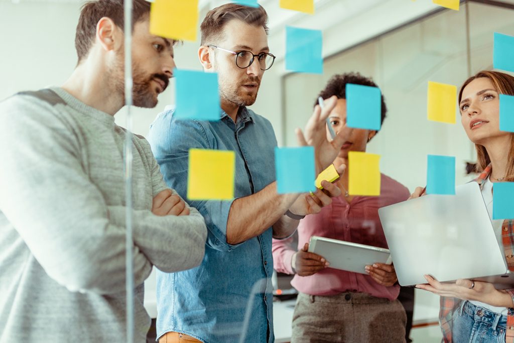 Vista a través del cristal de hombres y mujeres ejecutivos multiétnicos en ropa informal utilizando notas post it mientras comparten ideas en una oficina moderna.