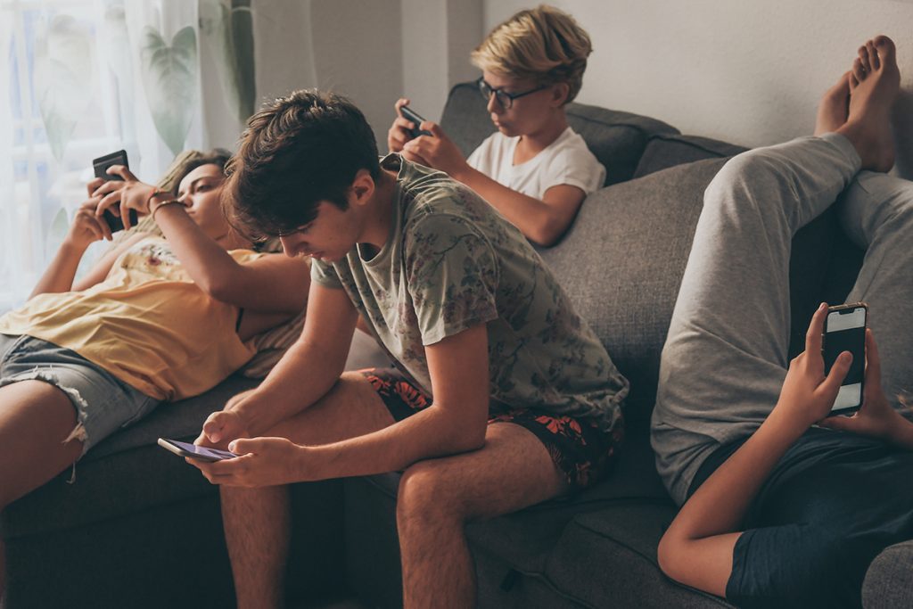 Grupo de adolescentes usando un smartphone sentados en el sofá de su casa. Chicos jóvenes y una chica compartiendo foto y vídeo viendo una historia social online. Amigos disfrutando de las nuevas tendencias tecnológicas. Concepto de juventud y tecnología