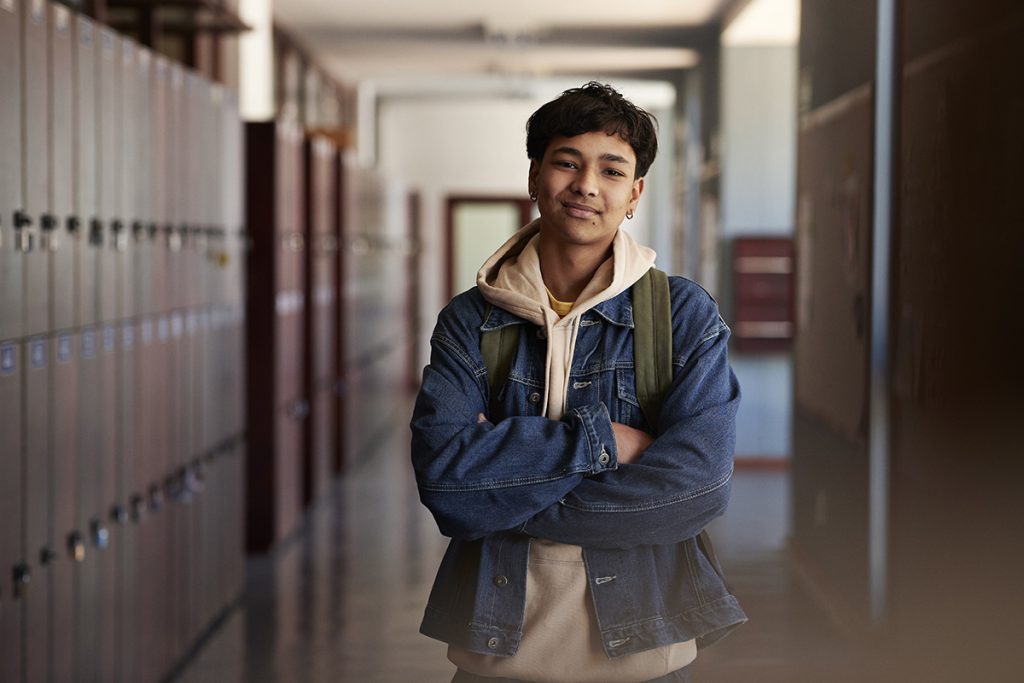 Retrato de adolescente sonriente con chaqueta vaquera de pie con los brazos cruzados en el pasillo del instituto